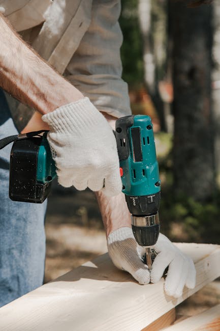 A construction worker using a cordless drill on a wooden panel outdoors, wearing protective gloves.